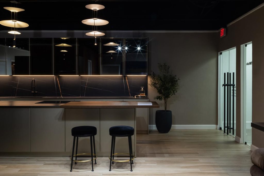 A modern kitchen showroom featuring a sleek design with a marble backsplash, under-cabinet lighting, a wooden countertop, and two black and gold stools. A potted plant adds a touch of greenery to the space.